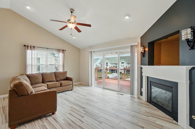 living area featuring baseboards, ceiling fan, a tiled fireplace, light wood finished floors, and vaulted ceiling