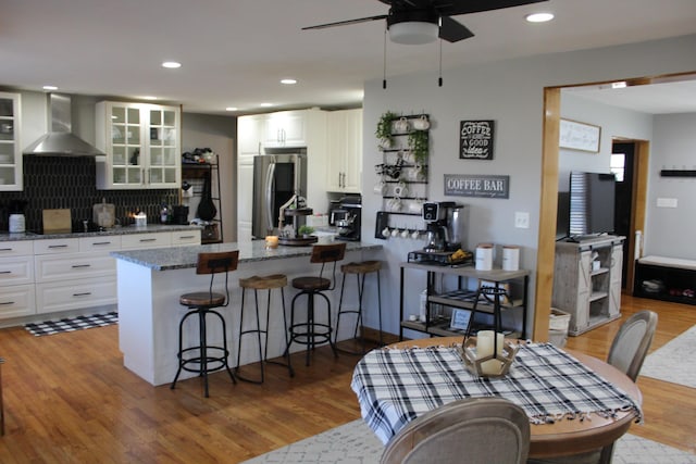 kitchen with glass insert cabinets, wall chimney range hood, a peninsula, smart refrigerator, and stone countertops
