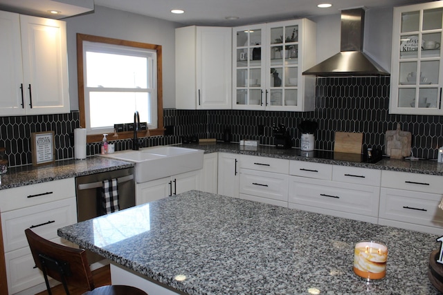 kitchen featuring a sink, wall chimney range hood, dark stone countertops, white cabinets, and stainless steel dishwasher