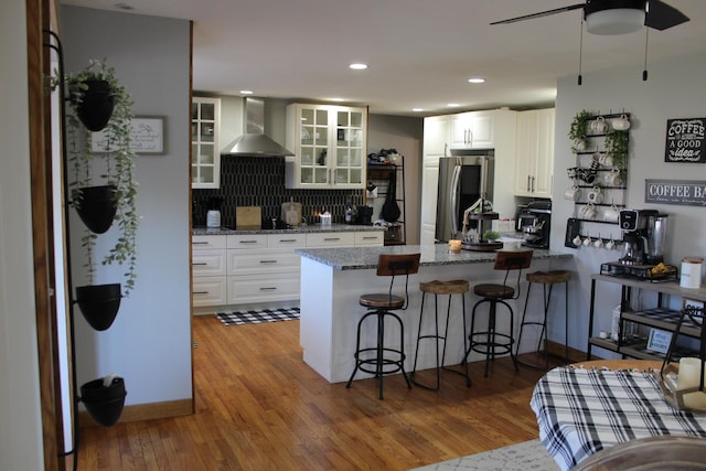 kitchen featuring glass insert cabinets, wall chimney range hood, a peninsula, smart refrigerator, and light wood-style floors