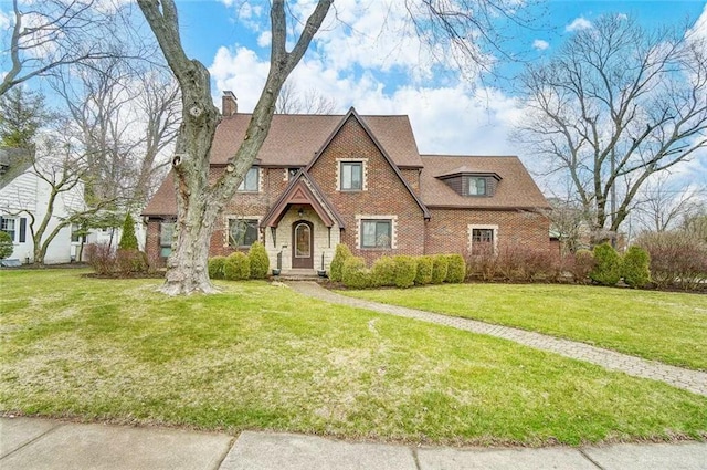 tudor home featuring brick siding, a chimney, a front lawn, and roof with shingles