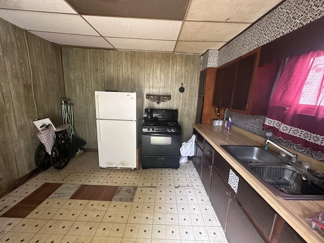 kitchen featuring sink, a drop ceiling, black range with gas stovetop, white refrigerator, and wooden walls