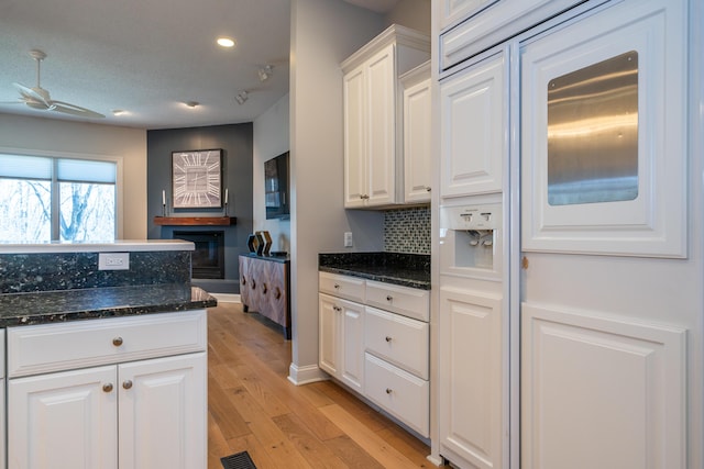 kitchen featuring ceiling fan, dark stone counters, light wood-type flooring, a glass covered fireplace, and white cabinetry