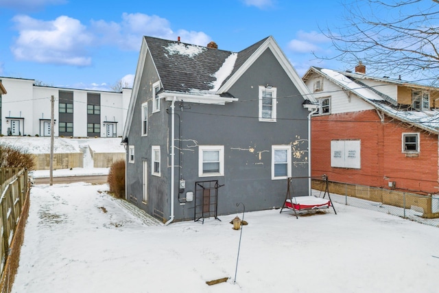 snow covered rear of property featuring roof with shingles, fence, and stucco siding