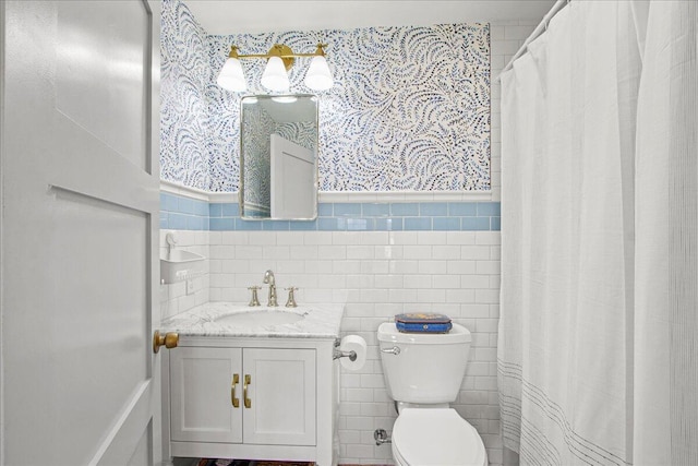 full bathroom featuring wallpapered walls, wainscoting, toilet, vanity, and tile walls