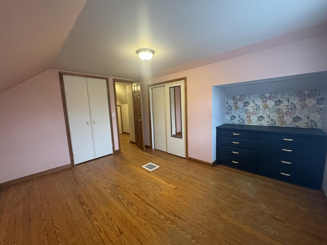 unfurnished bedroom with vaulted ceiling, wood finished floors, baseboards, visible vents, and two closets