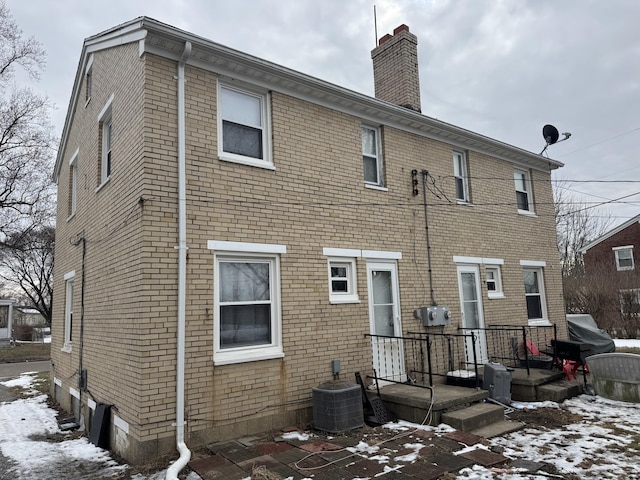 snow covered rear of property with central AC, brick siding, and a chimney