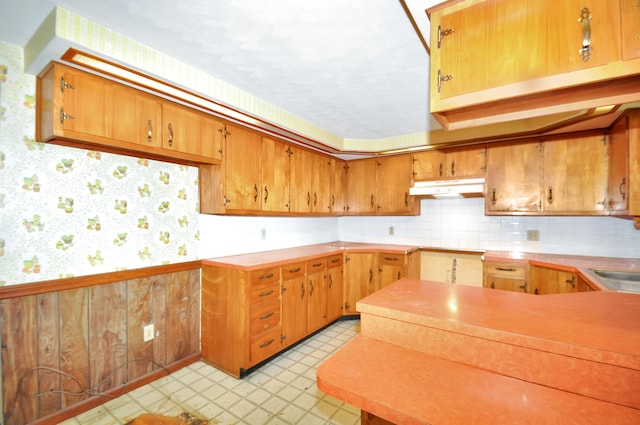 kitchen with light floors, wallpapered walls, under cabinet range hood, wainscoting, and brown cabinets