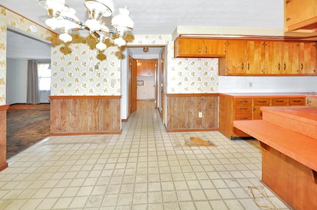 kitchen with wallpapered walls, a wainscoted wall, light countertops, an inviting chandelier, and brown cabinetry