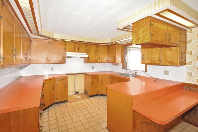 kitchen featuring a peninsula, a sink, under cabinet range hood, tasteful backsplash, and brown cabinets