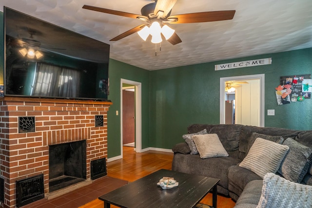 living room featuring a textured wall, a ceiling fan, a brick fireplace, wood finished floors, and baseboards