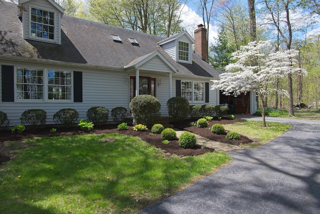 cape cod-style house featuring a shingled roof, aphalt driveway, a front yard, a chimney, and a garage