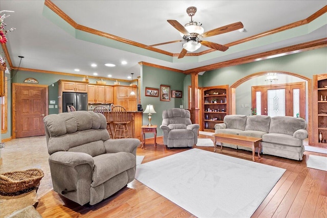 living room with arched walkways, a raised ceiling, light wood-style floors, ornamental molding, and baseboards