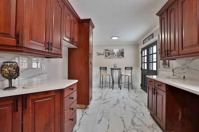 kitchen with backsplash, crown molding, light countertops, wainscoting, and marble finish floor