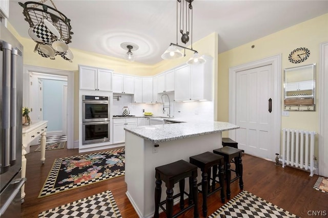 kitchen with stainless steel appliances, light stone countertops, decorative light fixtures, white cabinetry, and radiator heating unit