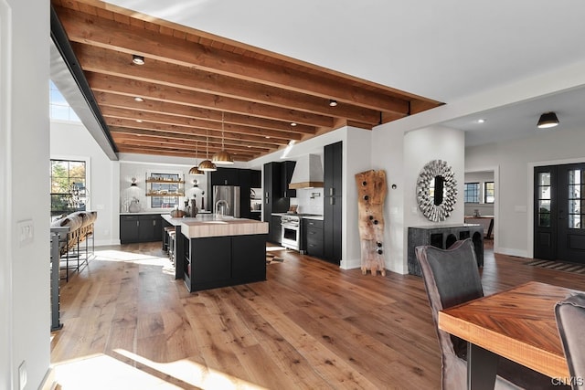 interior space featuring an island with sink, hardwood / wood-style floors, wall chimney range hood, hanging light fixtures, and beam ceiling