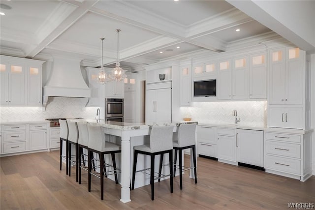 kitchen with coffered ceiling, appliances with stainless steel finishes, an island with sink, pendant lighting, and premium range hood