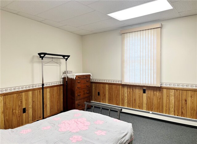 carpeted bedroom featuring wooden walls, a drop ceiling, and a baseboard heating unit