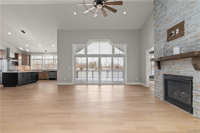 unfurnished living room with ceiling fan, a fireplace, a towering ceiling, sink, and light wood-type flooring