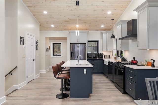 kitchen featuring an island with sink, wall chimney exhaust hood, black gas range, decorative light fixtures, and stainless steel refrigerator with ice dispenser