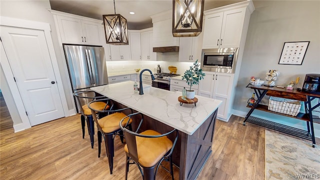 kitchen with stainless steel appliances, an island with sink, light stone counters, pendant lighting, and light wood-type flooring