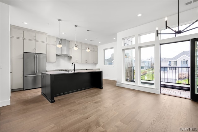 kitchen featuring high quality fridge, wood-type flooring, wall chimney exhaust hood, and an island with sink