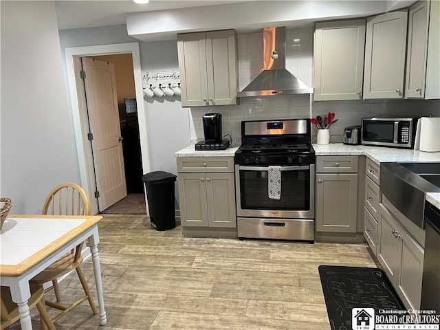 kitchen with wall chimney exhaust hood, gray cabinets, light hardwood / wood-style flooring, stainless steel appliances, and tasteful backsplash