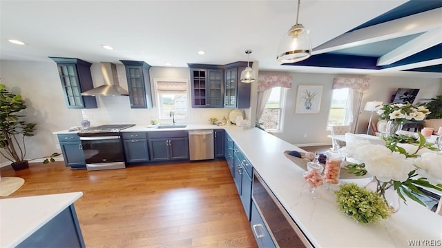 kitchen with light wood-type flooring, wall chimney exhaust hood, stainless steel appliances, sink, and pendant lighting