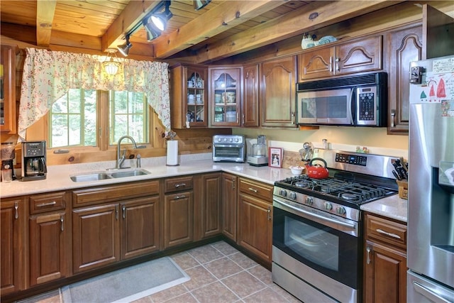 kitchen featuring a sink, light countertops, appliances with stainless steel finishes, beam ceiling, and glass insert cabinets