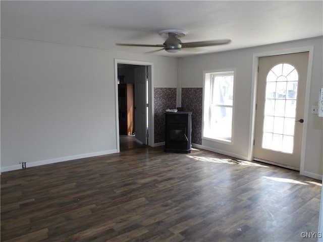 unfurnished living room featuring a wood stove, dark wood-style floors, baseboards, and ceiling fan