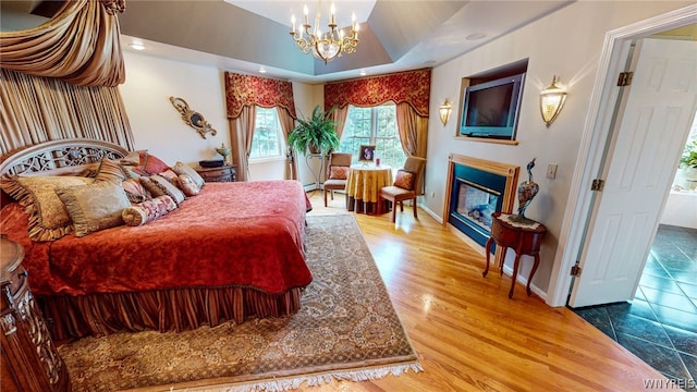 bedroom with a tray ceiling, a glass covered fireplace, wood finished floors, a chandelier, and baseboards