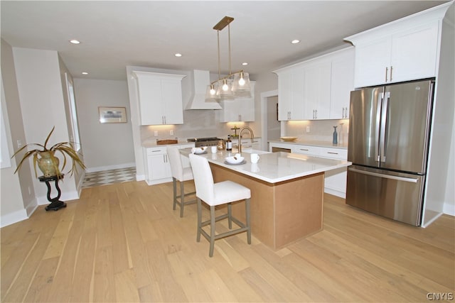 kitchen with stainless steel fridge, white cabinets, a center island with sink, premium range hood, and light wood-type flooring