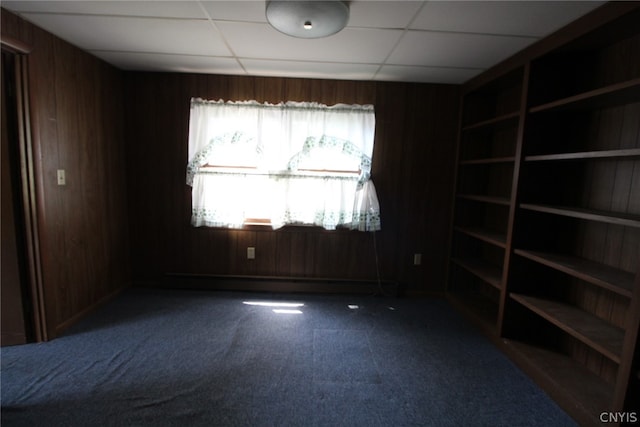 empty room with carpet flooring, a drop ceiling, wooden walls, and a baseboard radiator