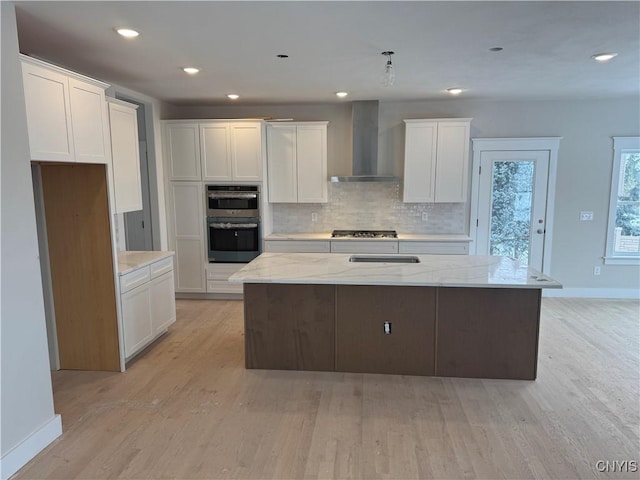 kitchen featuring white cabinetry, light stone countertops, wall chimney range hood, and a kitchen island with sink