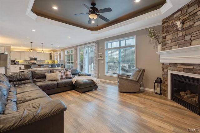 living room with crown molding, a stone fireplace, a raised ceiling, and light wood-type flooring