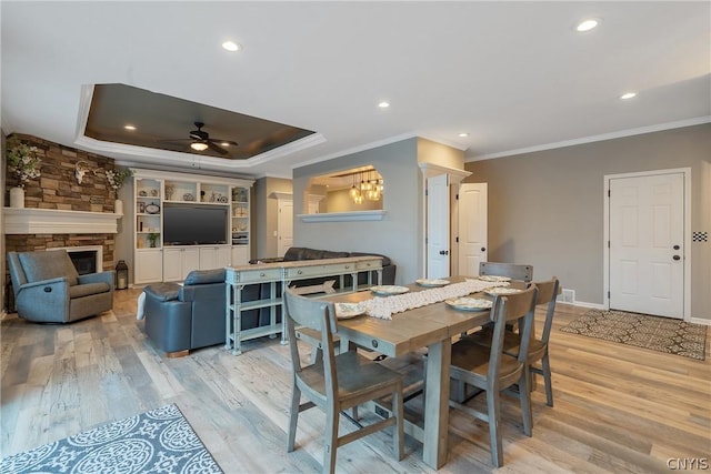 dining area featuring a stone fireplace, ceiling fan, a tray ceiling, crown molding, and light hardwood / wood-style flooring