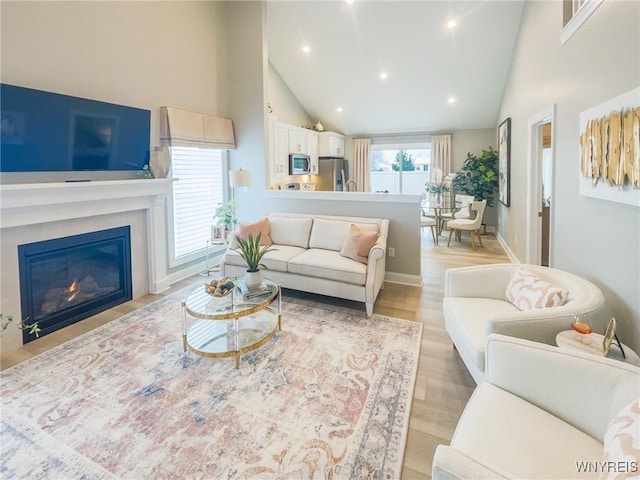 living area with light wood finished floors, recessed lighting, a glass covered fireplace, high vaulted ceiling, and baseboards