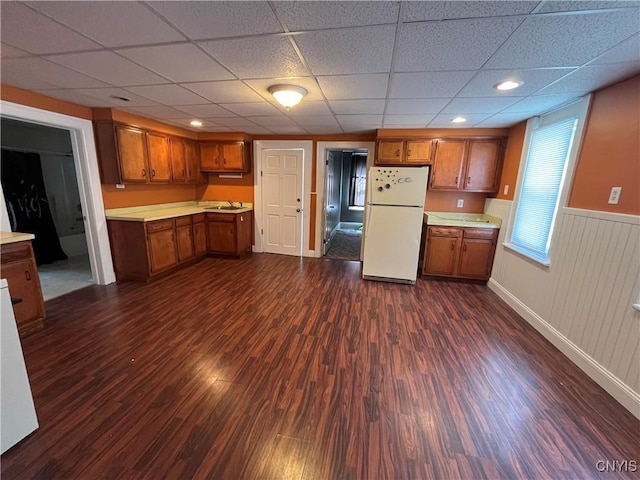 kitchen with a wainscoted wall, light countertops, freestanding refrigerator, brown cabinets, and dark wood-style floors