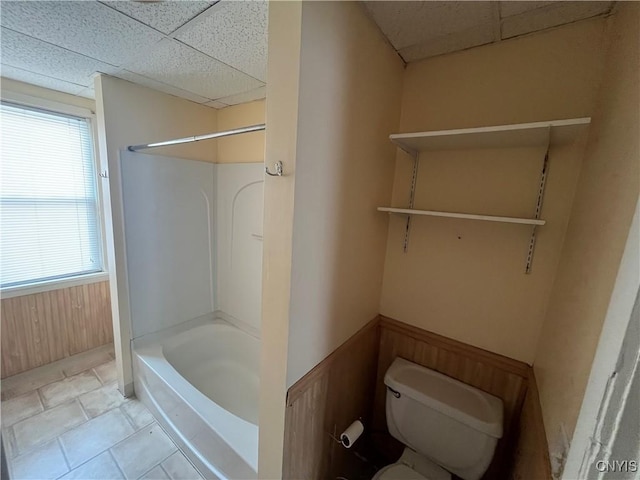 bathroom with a drop ceiling, tile patterned flooring, toilet, wooden walls, and a wainscoted wall