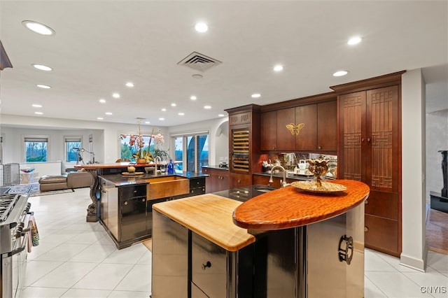 kitchen with a center island with sink, light tile patterned floors, butcher block counters, stainless steel stove, and dark brown cabinets