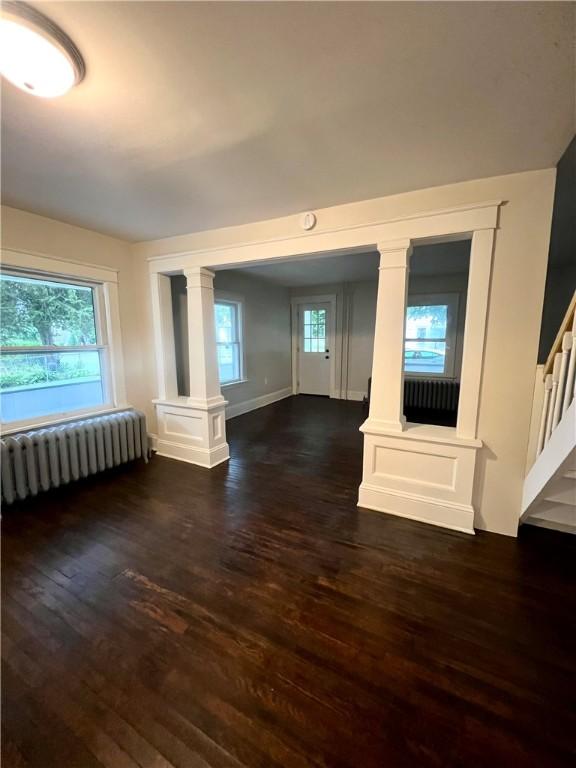 unfurnished living room featuring dark wood-type flooring, radiator, plenty of natural light, and ornate columns