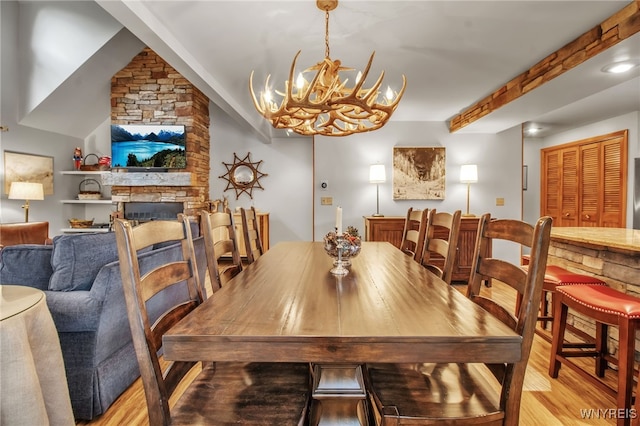 dining room with a notable chandelier, beam ceiling, light hardwood / wood-style floors, and a stone fireplace