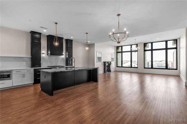 kitchen with backsplash, sink, a notable chandelier, hardwood / wood-style flooring, and a kitchen island with sink