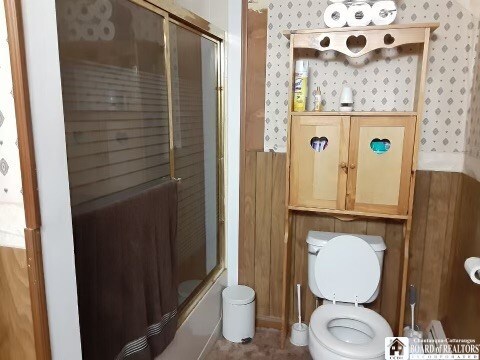 bathroom featuring wooden walls, toilet, and bath / shower combo with glass door