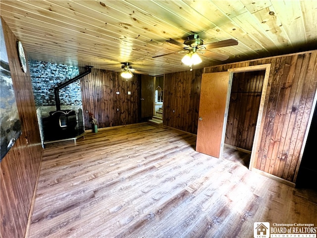 additional living space with wood walls, a wood stove, wooden ceiling, light wood-type flooring, and ceiling fan