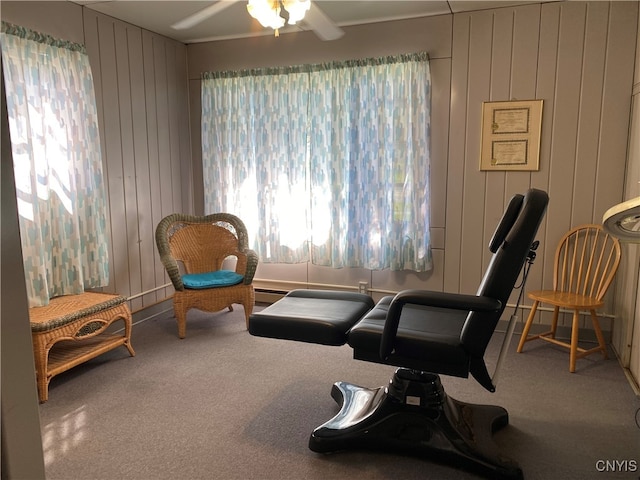 living area featuring a baseboard heating unit, wood walls, ceiling fan, and carpet flooring