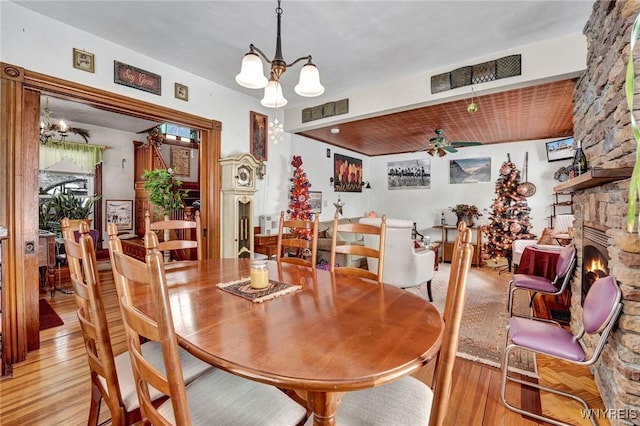 dining room with ceiling fan with notable chandelier, a stone fireplace, and light hardwood / wood-style flooring