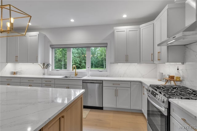 kitchen with light stone counters, light hardwood / wood-style floors, a notable chandelier, wall chimney exhaust hood, and stainless steel appliances