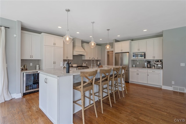 kitchen with custom range hood, stainless steel appliances, a kitchen island with sink, hardwood / wood-style flooring, and wine cooler