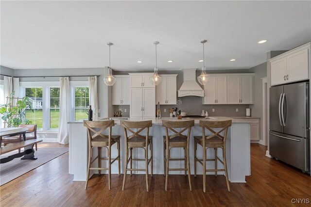 kitchen with stainless steel refrigerator, dark hardwood / wood-style floors, premium range hood, decorative light fixtures, and a kitchen island with sink